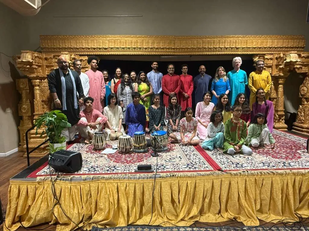 Musicians and students pose on a stage, with tabla drums on the rug in front