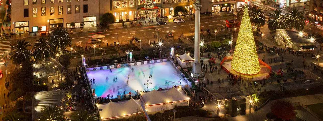 Union Square at night from above: the lit ice rink, the tall Christmas tree and the monument column beside it, with shops and streets all around