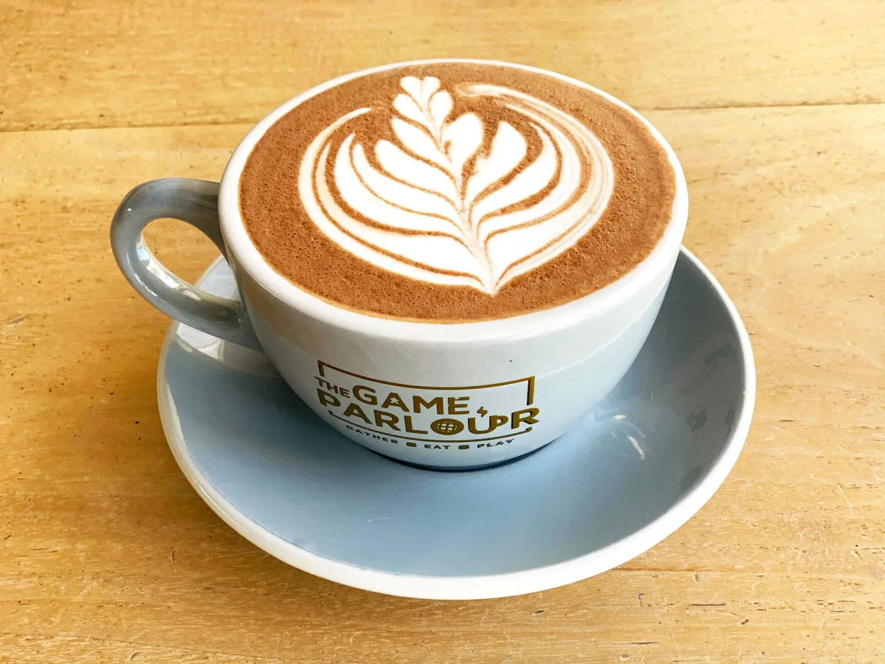 木桌上一杯拉花热巧克力，杯身印着 THE GAME PARLOUR 标志