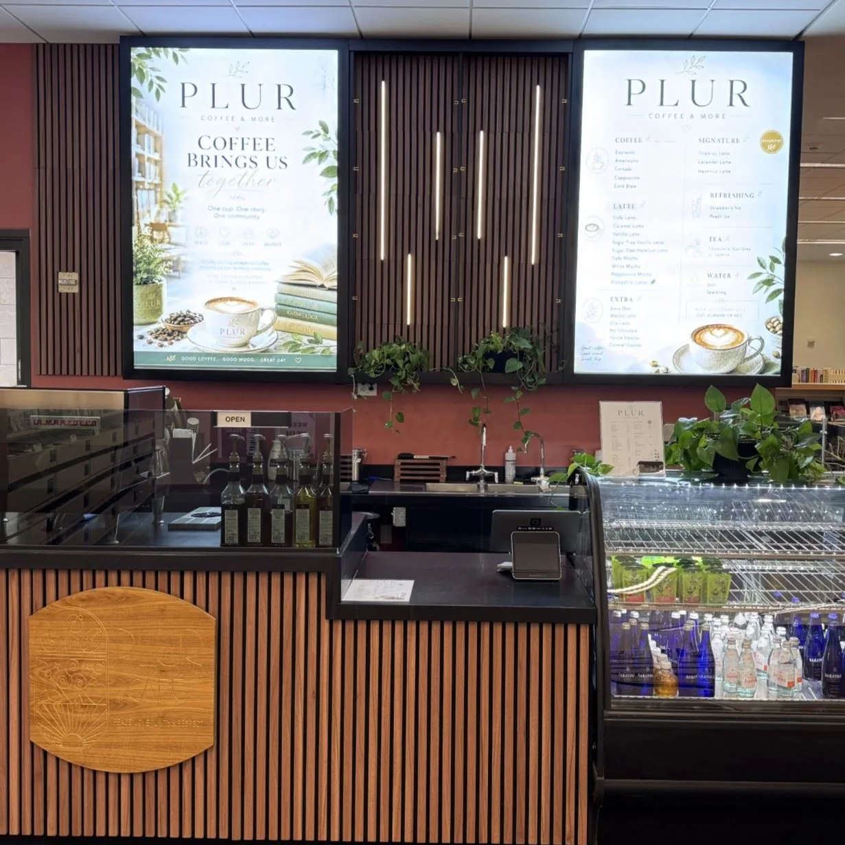图书馆里的 PLUR 咖啡吧台：两块 PLUR 菜单灯箱、木条吧台和冷藏饮料柜